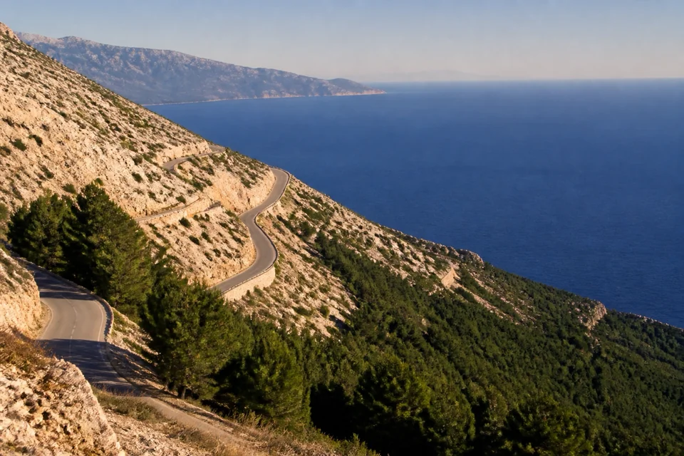 A winding mountain road dropping through pine slopes to deep blue water in Albania