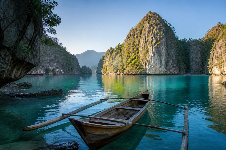 A wooden boat floating on still turquoise water among steep limestone islets in the Philippines