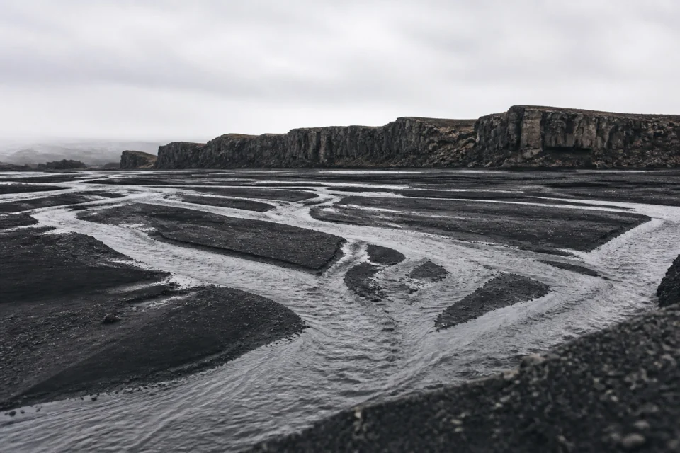A glacial stream braiding across a black sand plain before basalt cliffs in Iceland
