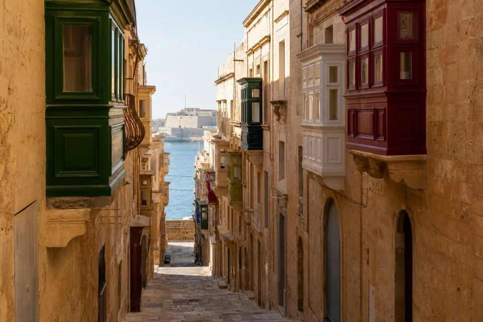 The harbour seen between green and maroon wooden balconies on a narrow Malta street
