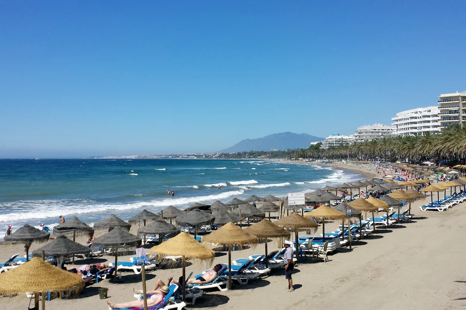 A beach with straw umbrellas, waterfront buildings and mountains behind in Marbella